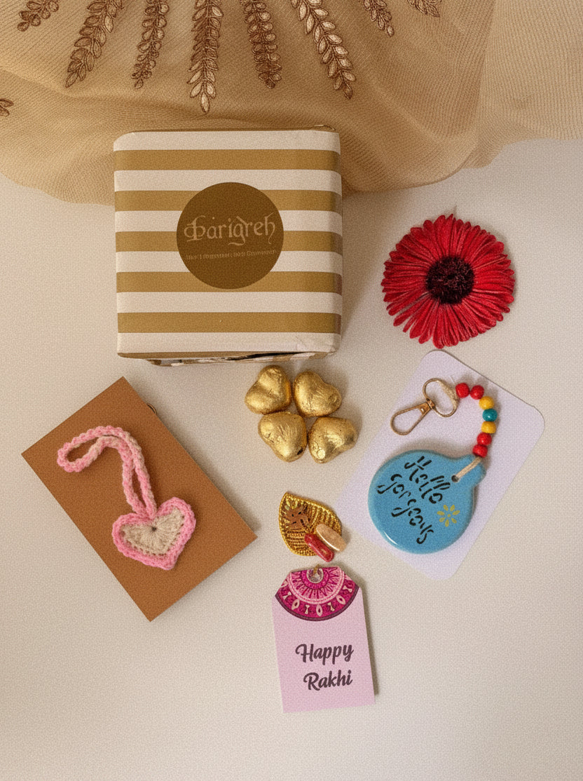 A styled flat-lay of a Rakhi gift set on a cream background. The set includes a striped paper Rakhi box with a gold logo, a pink and gold embroidered heart-shaped Rakhi, three gold foil heart candies, a red flower embellishment, a blue 'hello for you' card, and a pink 'Happy Rakhi' card.