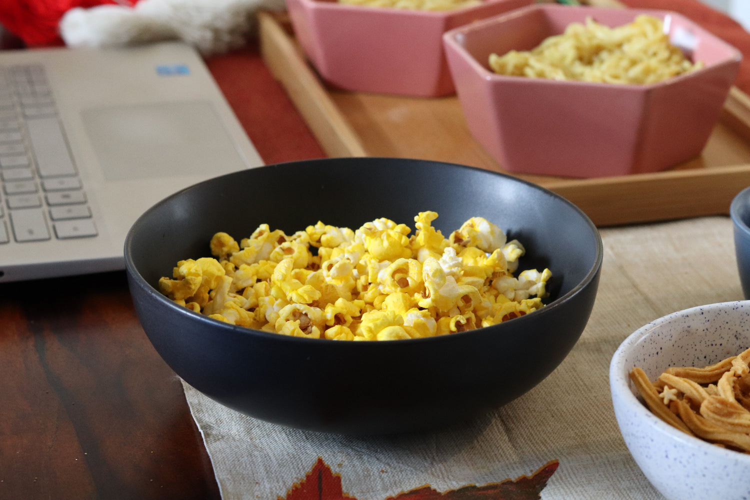 Black bowl of popcorn on a table with a laptop and other bowls in the background