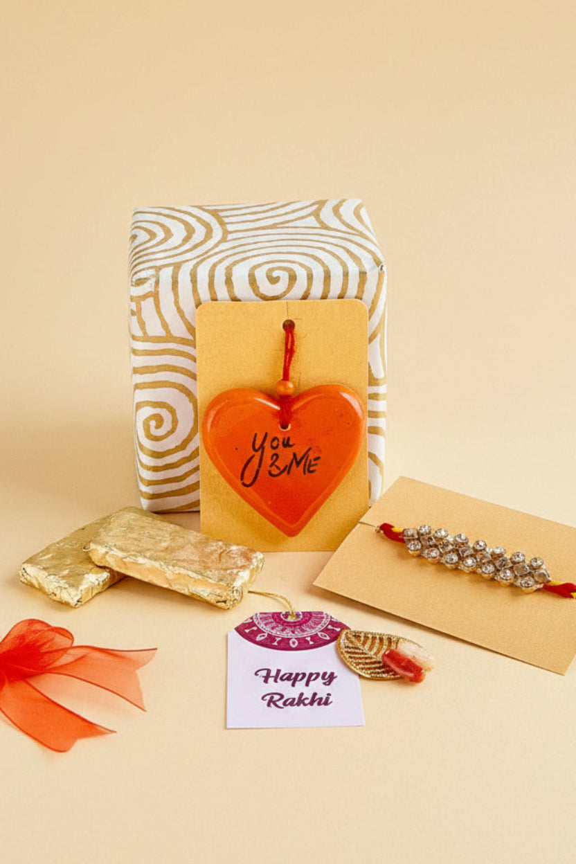 Gift box with heart-shaped charm hanging with you & me text on it, Rakhi, homemade chocolates and 'Happy Rakhi' card.